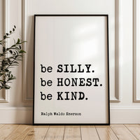 A framed quote by Ralph Waldo Emerson, "be SILLY. be Honest. be KIND.", is displayed on a wooden floor against a white wall.