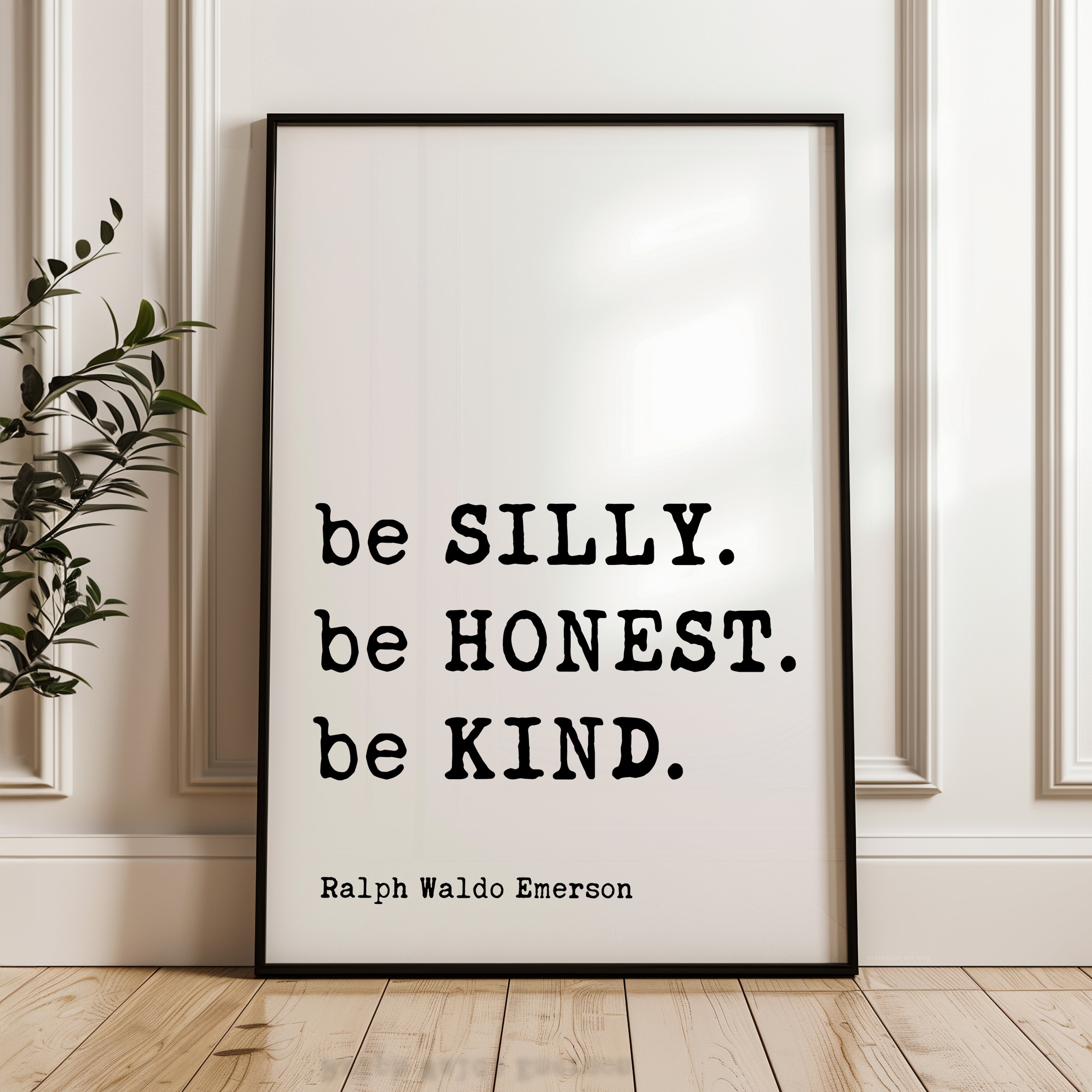 A framed quote by Ralph Waldo Emerson, "be SILLY. be Honest. be KIND.", is displayed on a wooden floor against a white wall.