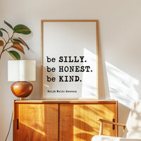 a framed quote by Ralph Waldo Emerson, "be SILY. be HONEST. be KIND.", placed on a wooden cabinet or dresser in a room with a lamp and a plant.