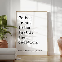 a quote by William Shakespeare, "To be, or not to be: that is the question." The quote is displayed on a white poster or sign, which is placed on a wall in a room with a potted plant and a chair.