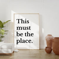 a large white poster with the text "This must be the place" in bold black letters, placed in a room with a white wall and a wooden floor.