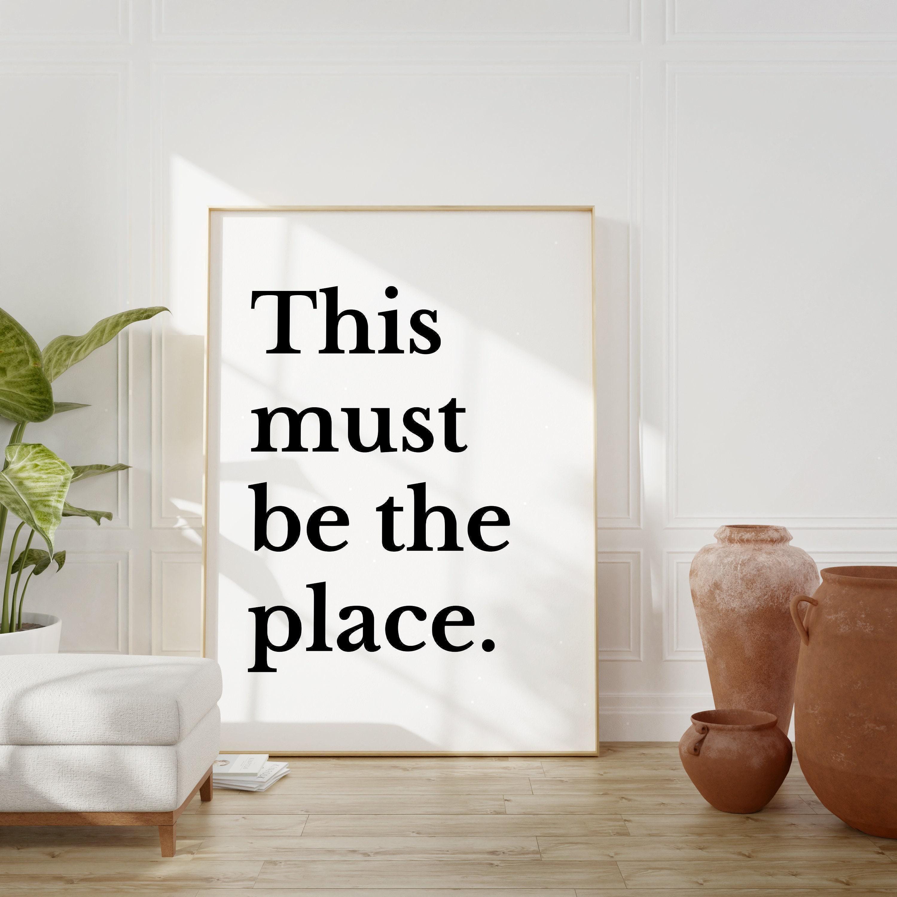 a large white poster with the text "This must be the place" in bold black letters, placed in a room with a white wall and a wooden floor.