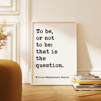 a quote by William Shakespeare, "To be, or not to be: that is the question." The quote is displayed on a white poster, which is propped up against a white wall. In the foreground, there is a stack of books and a plant.