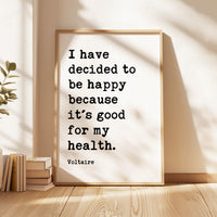 a framed quote by Voltaire, "I have decided to be happy because it's good for my health," placed on a wooden floor next to a stack of books.