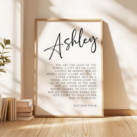 May include: A black and white printable wall art design with the name Ashley at the top. The text reads: 'You are the light of the world. A city set on a hill cannot be hidden. Nor do people light a lamp and put it under a basket, but on a stand, and it gives light to all in the house. In the same way, let your light shine before others, so that they may see your good works and give glory to your Father who is in heaven. Matthew 5:14-16'