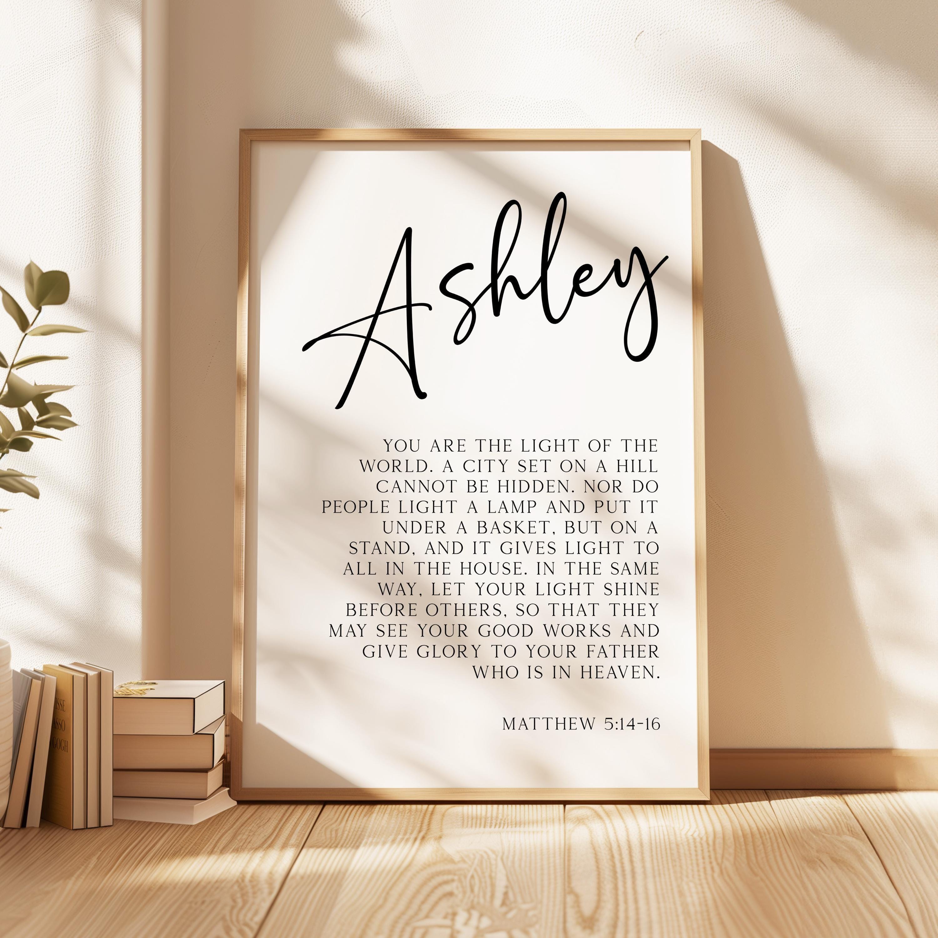 May include: A black and white printable wall art design with the name Ashley at the top. The text reads: 'You are the light of the world. A city set on a hill cannot be hidden. Nor do people light a lamp and put it under a basket, but on a stand, and it gives light to all in the house. In the same way, let your light shine before others, so that they may see your good works and give glory to your Father who is in heaven. Matthew 5:14-16'