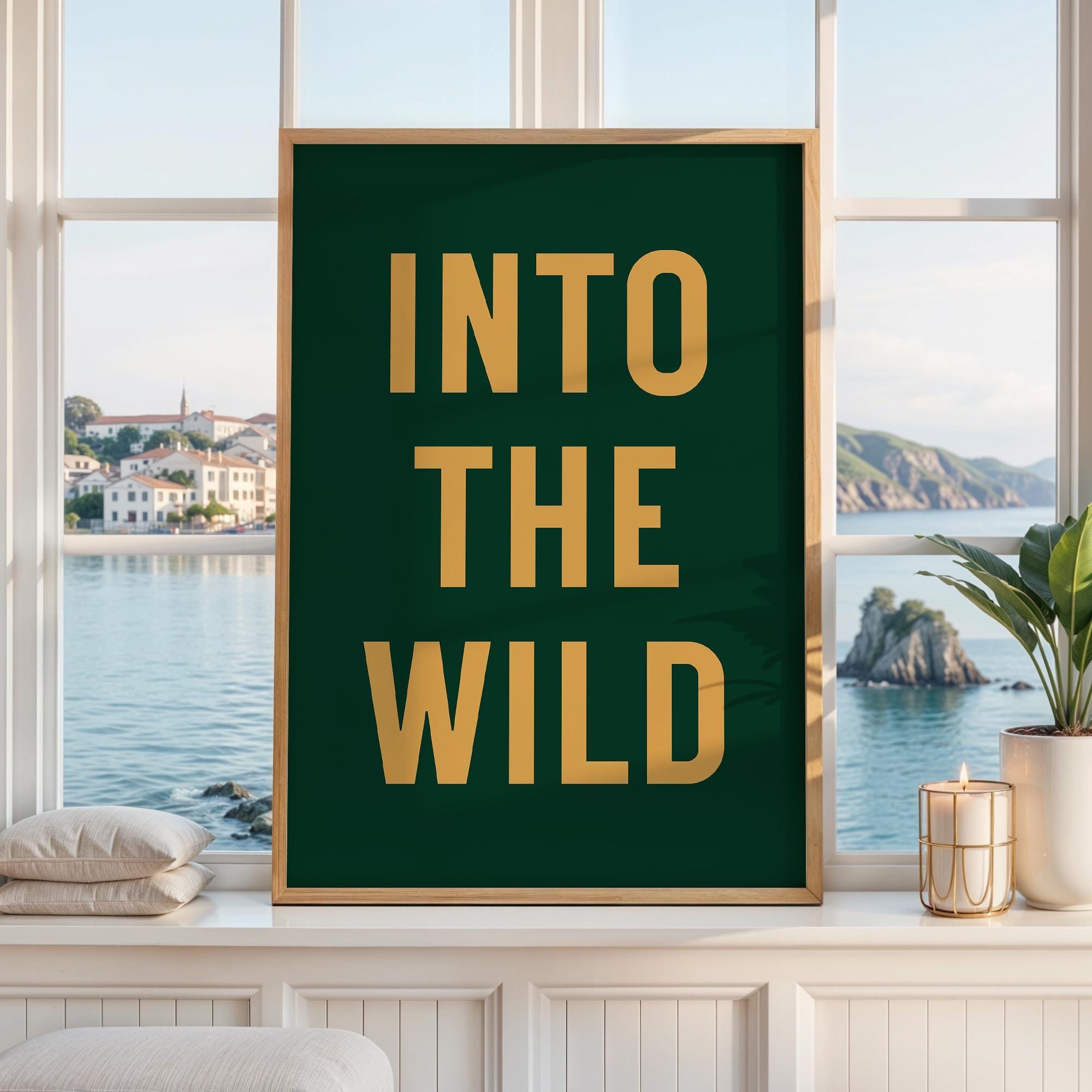 May include: A rectangular wall art print in a light wood frame featuring a dark forest green background with the text INTO THE WILD printed in bold, gold-colored capital letters. The artwork is displayed against a window with a coastal view.