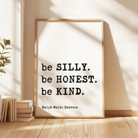 A framed quote by Ralph Waldo Emerson, "be SILLY. be HONEST. be KIND.", is displayed on a wooden floor next to a stack of books.