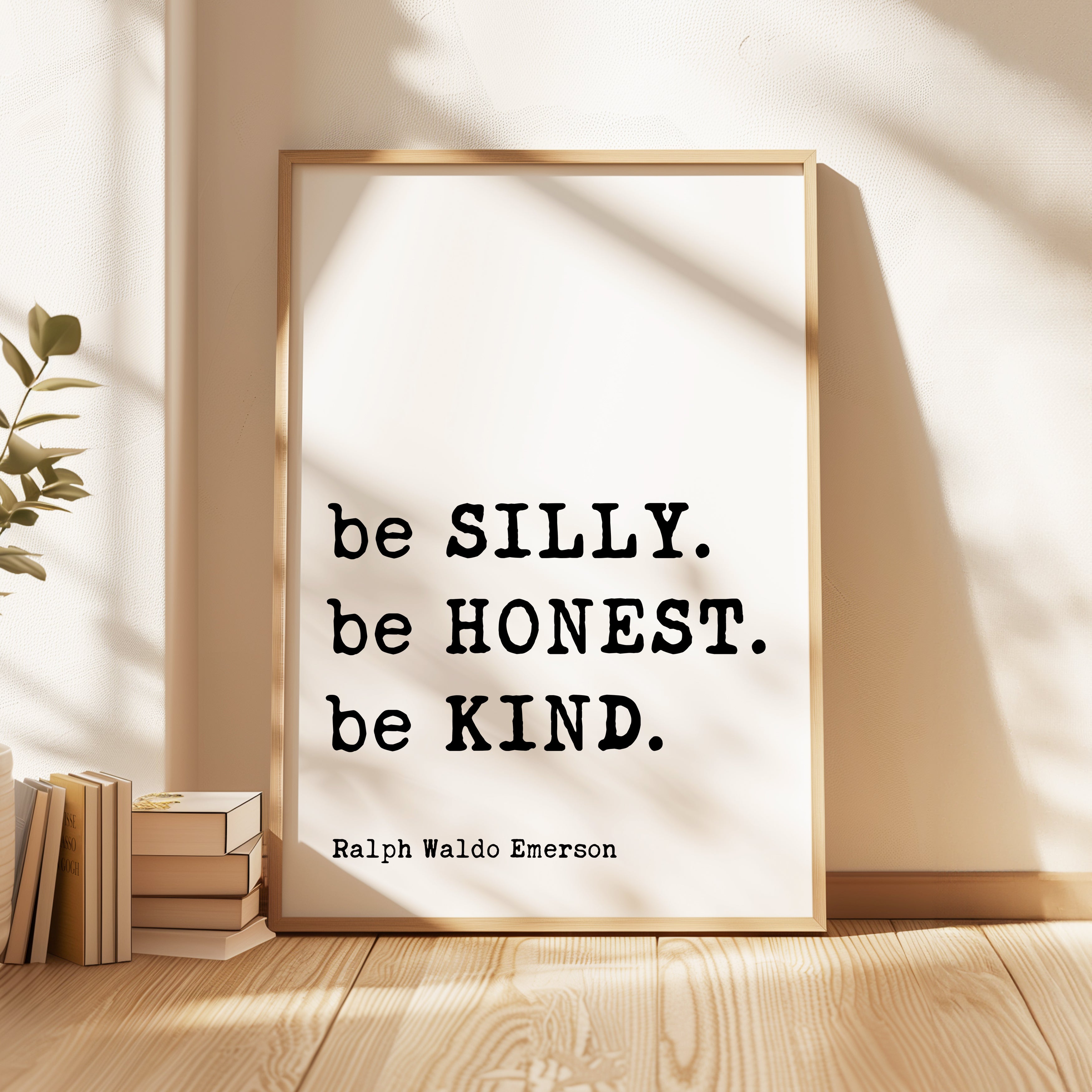 A framed quote by Ralph Waldo Emerson, "be SILLY. be HONEST. be KIND.", is displayed on a wooden floor next to a stack of books.