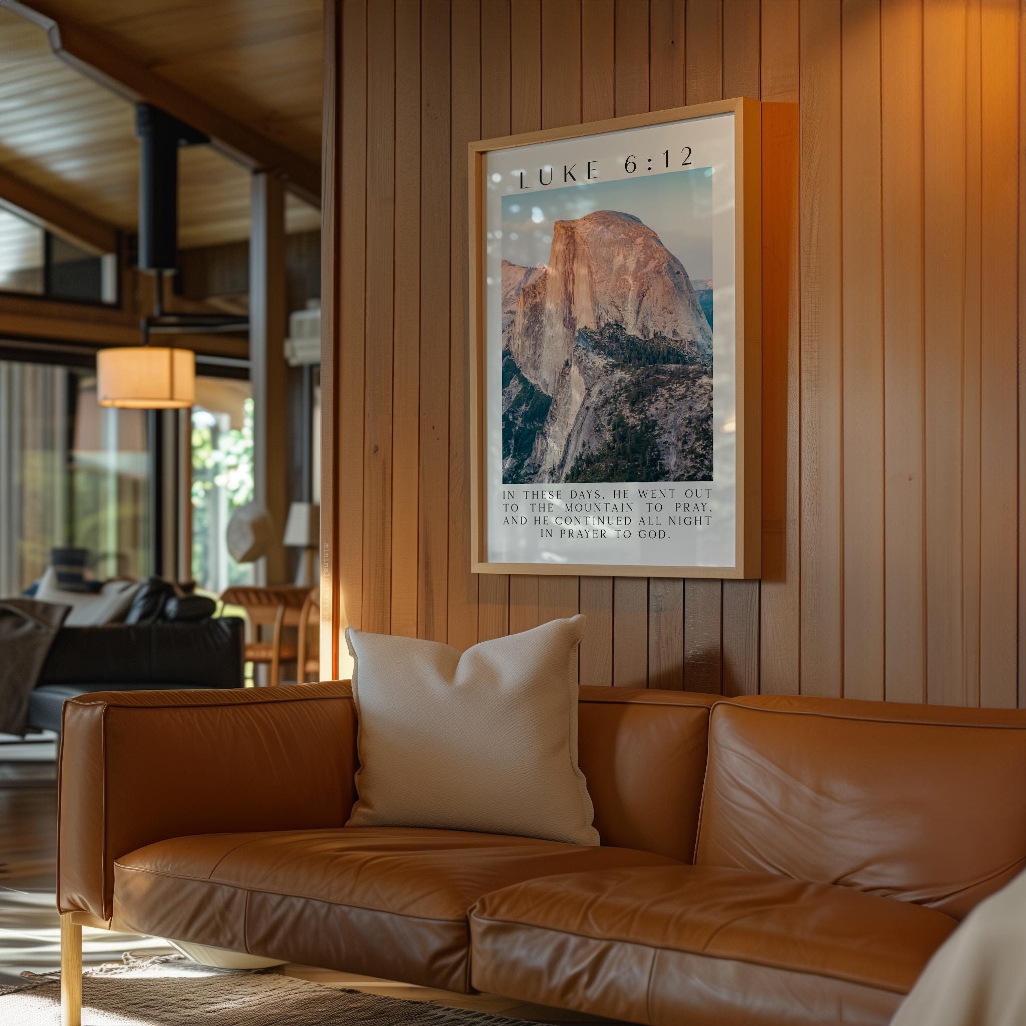 a large framed picture of a mountain, hanging on a wooden wall in a room with a brown leather couch and a window.