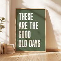 May include: A framed art print with the words "THESE ARE THE GOOD OLD DAYS" in bold white letters on a dark green background. The frame is a light brown color. The print is leaning against a wall on a wooden floor.