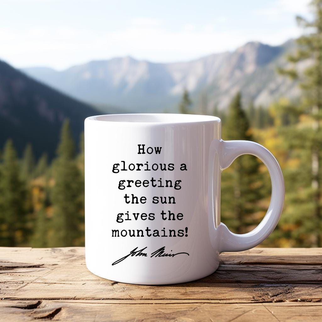 May include: White ceramic coffee mug with a black quote that reads 'How glorious a greeting the sun gives the mountains!' with the signature 'John Muir' underneath. The mug is sitting on a wooden surface.
