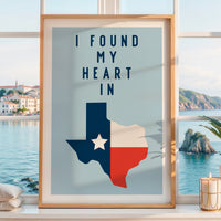 a framed poster with a Texas state map and the text "I FOUND MY HEART IN" displayed on it, placed on a window sill overlooking a scenic coastal view.