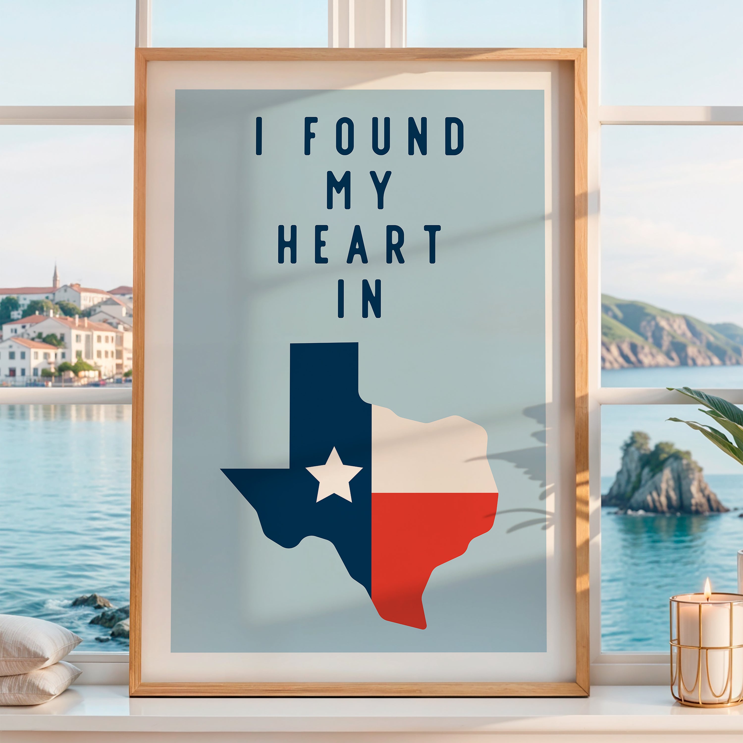 a framed poster with a Texas state map and the text "I FOUND MY HEART IN" displayed on it, placed on a window sill overlooking a scenic coastal view.