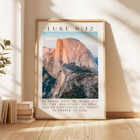 A framed poster of a mountain landscape with a Bible verse from Luke 6:12 displayed on it, placed on a wooden floor next to a stack of books.