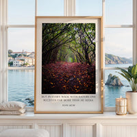 A framed photograph of a forest path with fallen leaves, displayed on a white shelf in a room with a view of a body of water and a distant town.