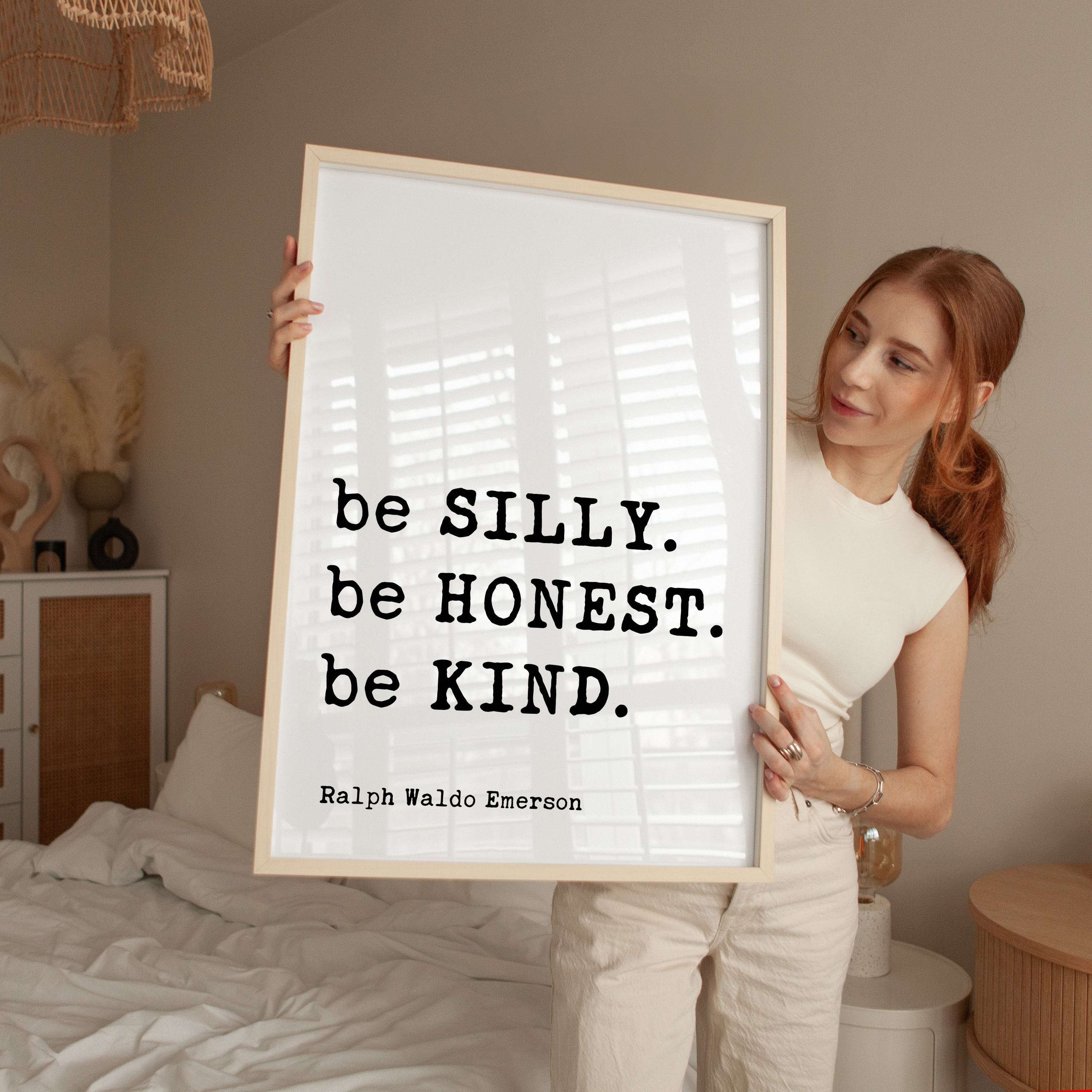 A woman holding a framed poster with the text "be SILLY. be HONEST. be KIND." by Ralph Waldo Emerson.