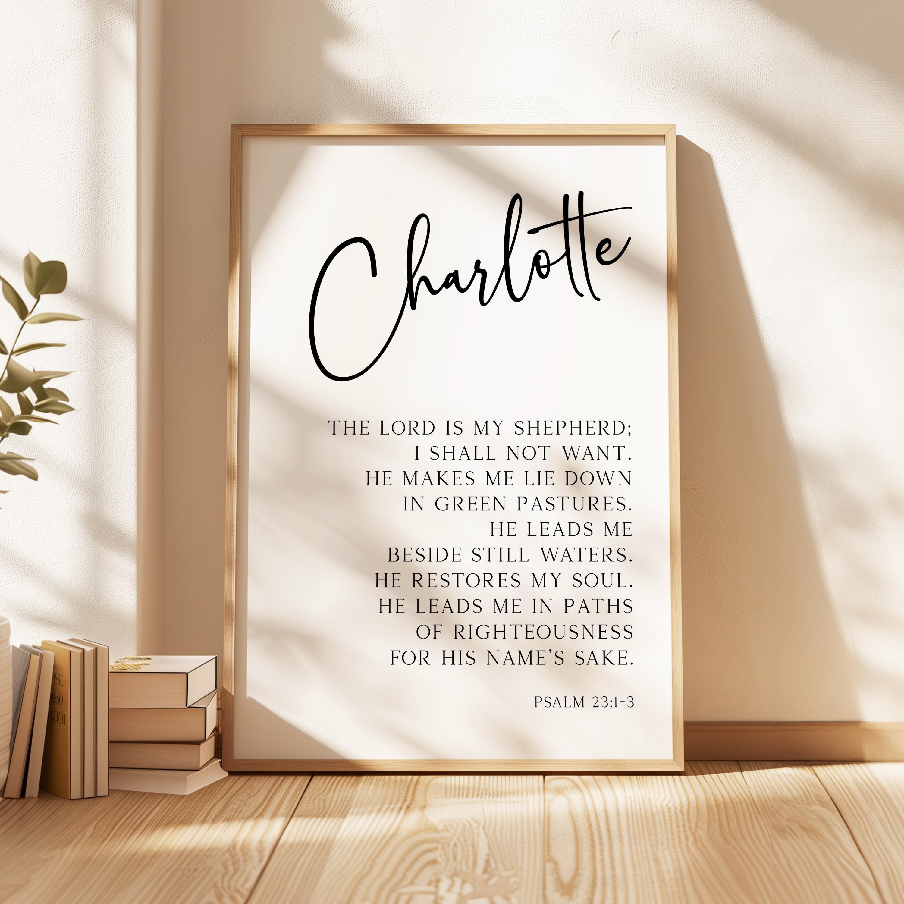 a framed Bible verse titled "Charlotte" with a black cursive font, placed on a wooden floor next to a stack of books.