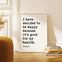 a framed quote by Voltaire, "I have decided to be happy because it's good for my health," placed on a wooden floor next to a stack of books.