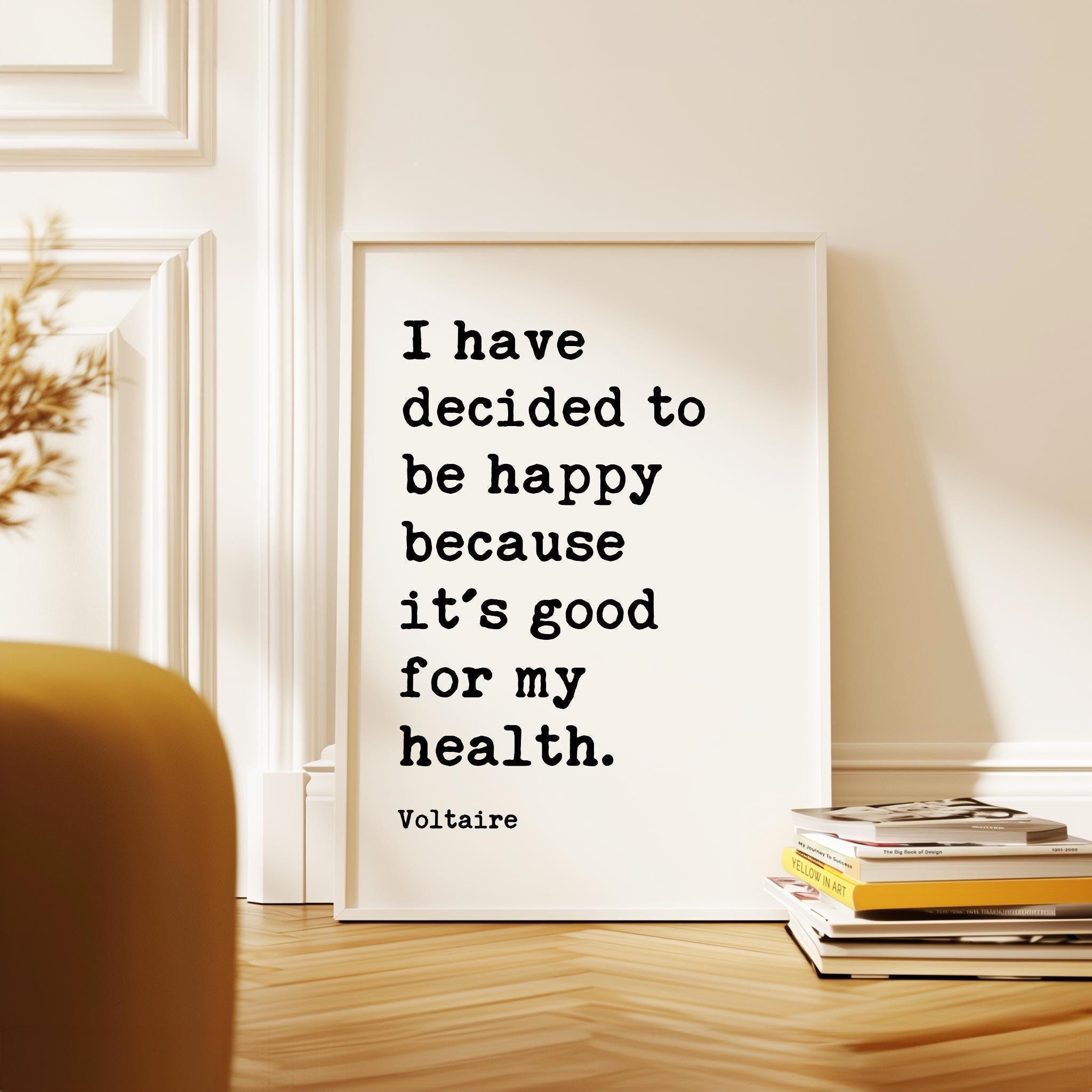 a framed quote by Voltaire, "I have decided to be happy because it's good for my health," placed on a wooden floor next to a stack of books.