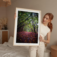 A woman holds a framed photograph of a forest path with fallen leaves, standing in a bedroom with a bed and other furniture visible in the background.