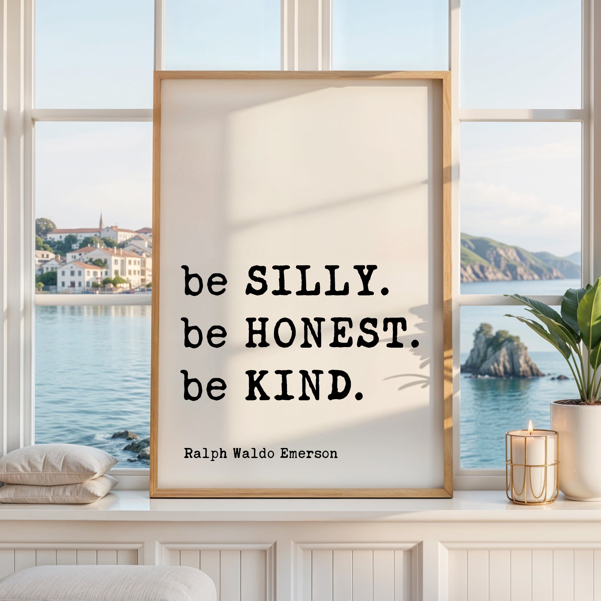 a framed quote by Ralph Waldo Emerson, "be SILLY. be Honest. be KIND.", placed on a window sill with a view of a coastal landscape.