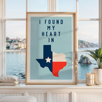 a framed poster with a Texas state map and the text "I FOUND MY HEART IN" displayed on it, placed on a window sill with a view of the ocean and mountains.