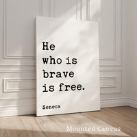 A white canvas with the quote "He who is brave is free" by Seneca, standing on a wooden floor against a white wall.