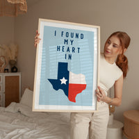 A woman holding a framed poster with a Texas state map and the text "I FOUND MY HEART IN"