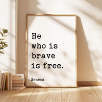 a framed quote by Seneca, "He who is brave is free", placed on a wooden floor next to a stack of books.