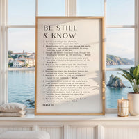 May include: A framed print with the words "BE STILL & KNOW" in black text. The print includes a passage from Psalm 46. The frame is a light brown color. A candle and a plant are visible in the background.