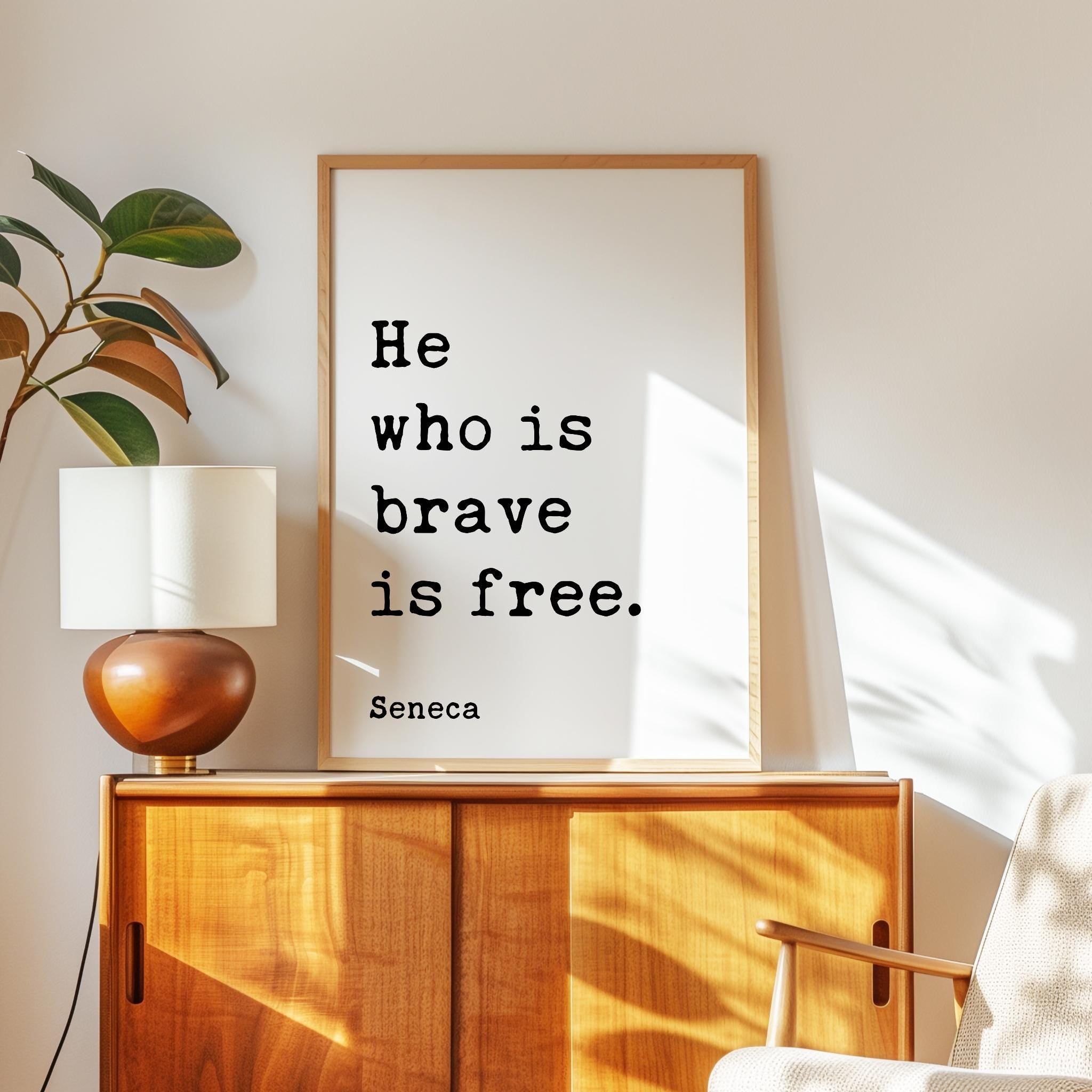 a framed quote by Seneca, "He who is brave is free," placed on a wooden cabinet or dresser in a room with a lamp and a plant.