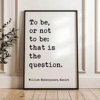 A framed quote by William Shakespeare, "To be, or not to be: that is the question," is displayed on a wooden floor against a white wall.