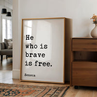 a framed quote by Seneca, "He who is brave is free." The quote is displayed on a white background, and the frame is placed on a wooden floor next to a wooden dresser.