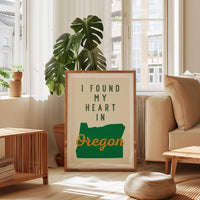 a framed poster with the text "I FOUND MY HEART IN OREGON" displayed prominently in the center. The poster is placed in a living room setting, with a couch, a coffee table, and a potted plant visible in the background.
