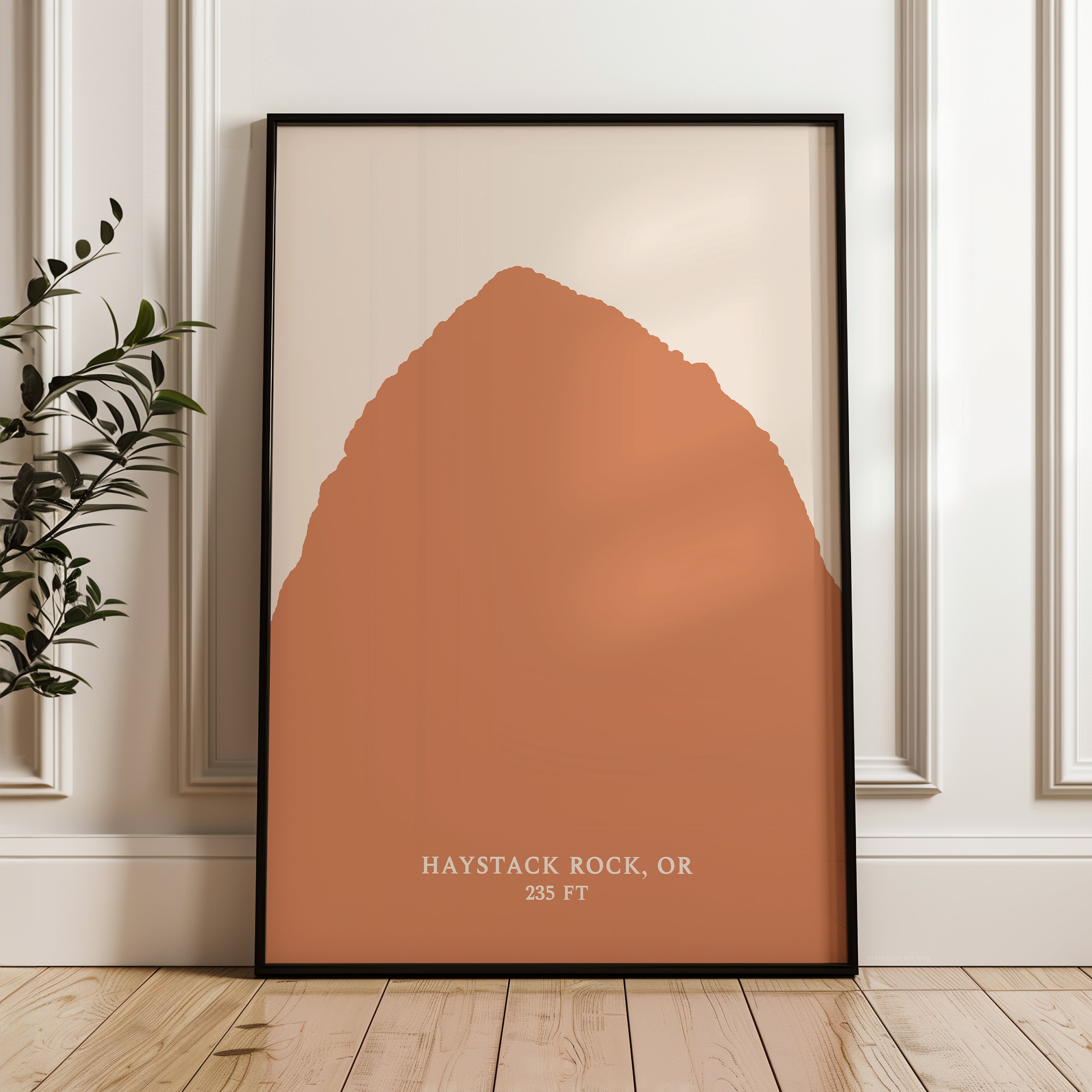 A framed poster of a mountain, with the text "HAYSTACK ROCK, OR" and "235 FT" printed on it, is displayed on a wooden floor next to a plant.