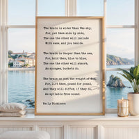 May include: A framed print featuring a poem by Emily Dickinson. The text is in black against a cream-colored background. The frame is a light brown color. The print is displayed on a white windowsill with a view of the ocean.