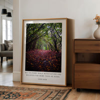 A framed photograph of a forest path with fallen leaves is displayed on a wall in a living room, accompanied by a quote attributed to John Muir.
