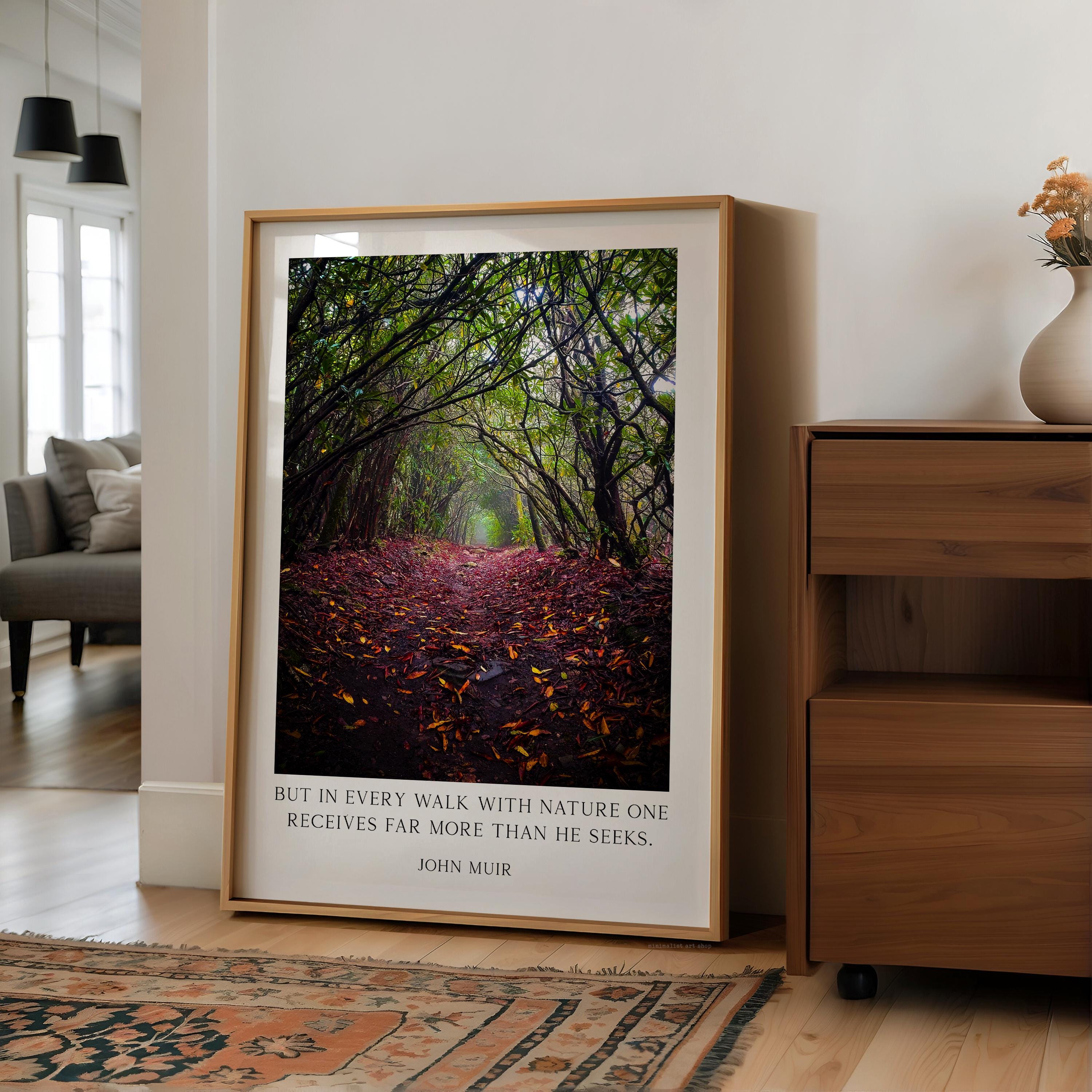 A framed photograph of a forest path with fallen leaves is displayed on a wall in a living room, accompanied by a quote attributed to John Muir.