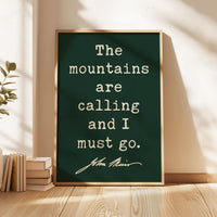 May include: A framed print with a dark green background and gold frame. The text reads "The mountains are calling and I must go." The quote is attributed to John Muir. The print is displayed on a wooden floor.