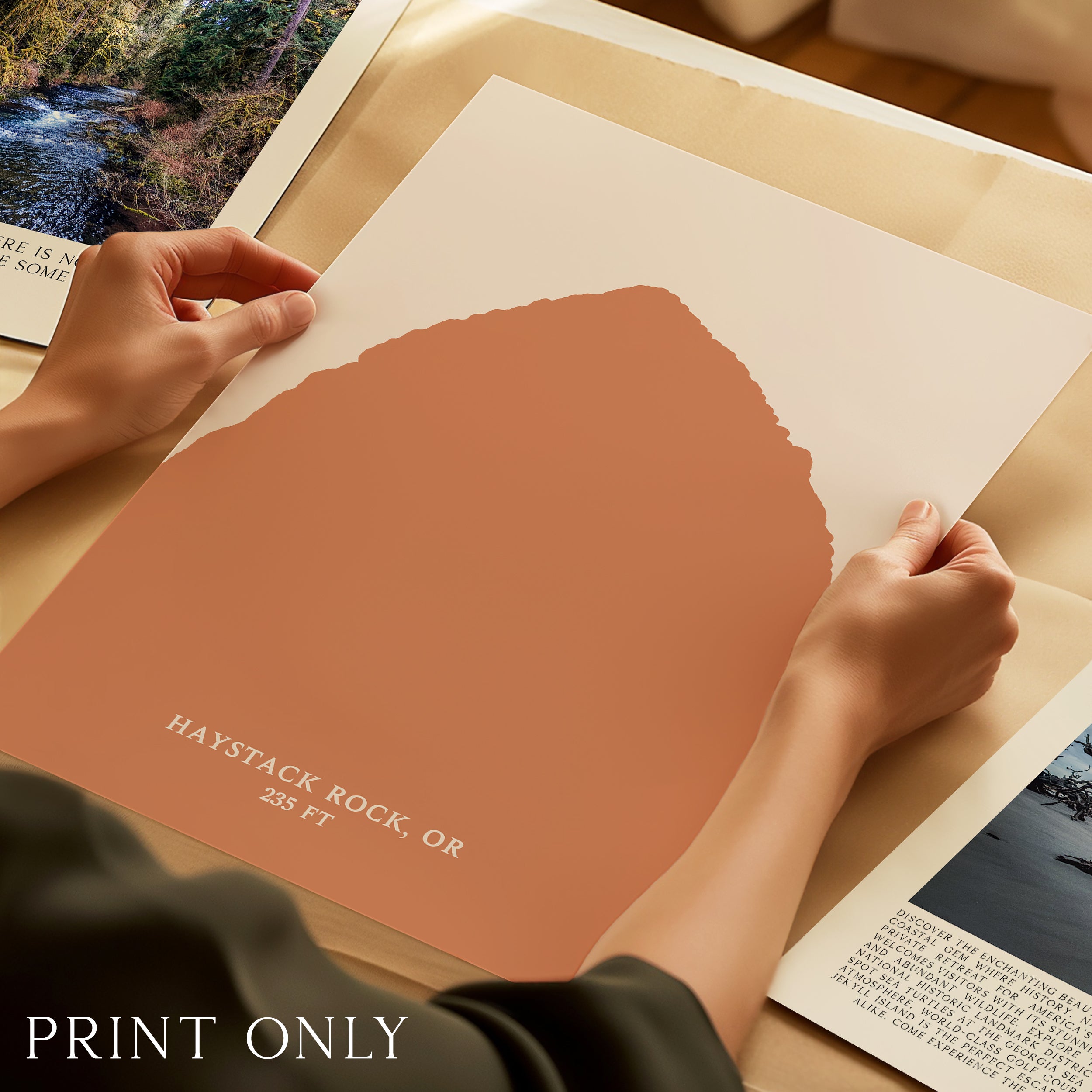 A person's hands are holding a large piece of paper with a printed image of a mountain range, and the text "HAYSTACK ROCK, OR" is visible.