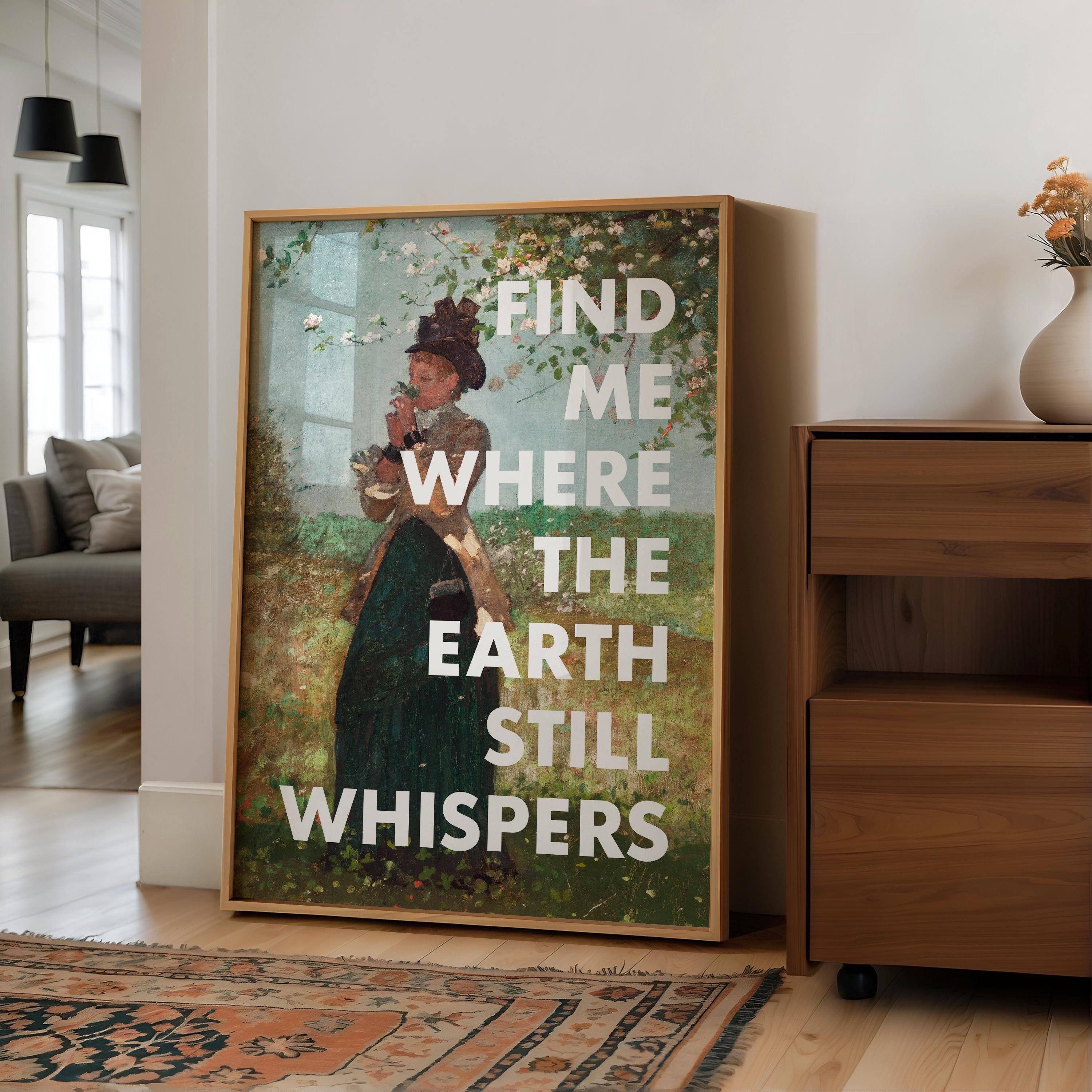 May include: A framed art print featuring a vintage-style painting of a woman in a garden, with the text "FIND ME WHERE THE EARTH STILL WHISPERS" in white. The artwork is in a wooden frame, displayed in a well-lit room.