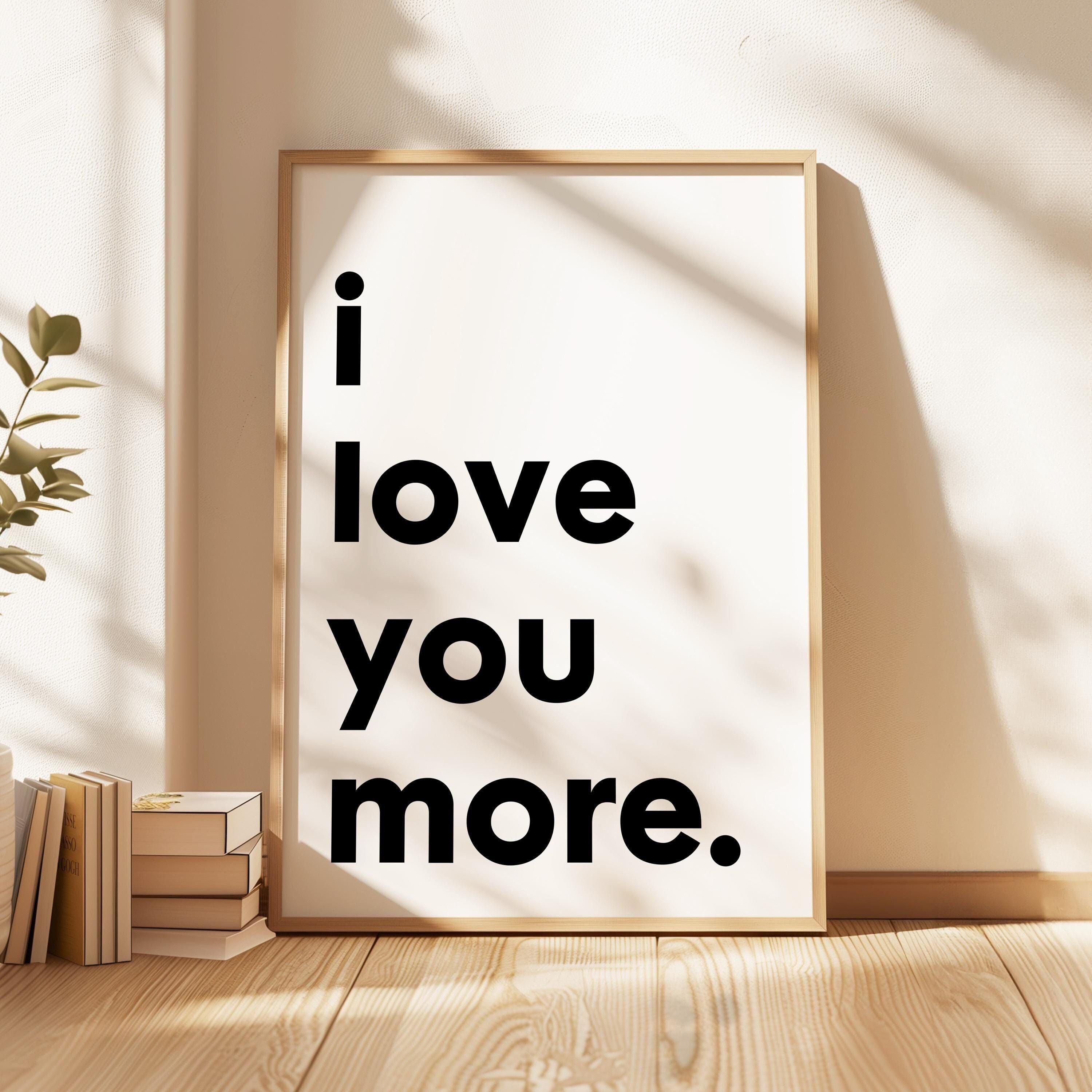 May include: A minimalist poster with a beige wooden frame features the text 'i love you more.' in bold, black, sans-serif font on a white background. The text is vertically stacked, with each word on a separate line. The poster is displayed against a light beige wall.