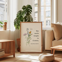 a framed quote by Ralph Waldo Emerson, "The earth laughs in flowers," placed in a living room setting with a potted plant and a window.