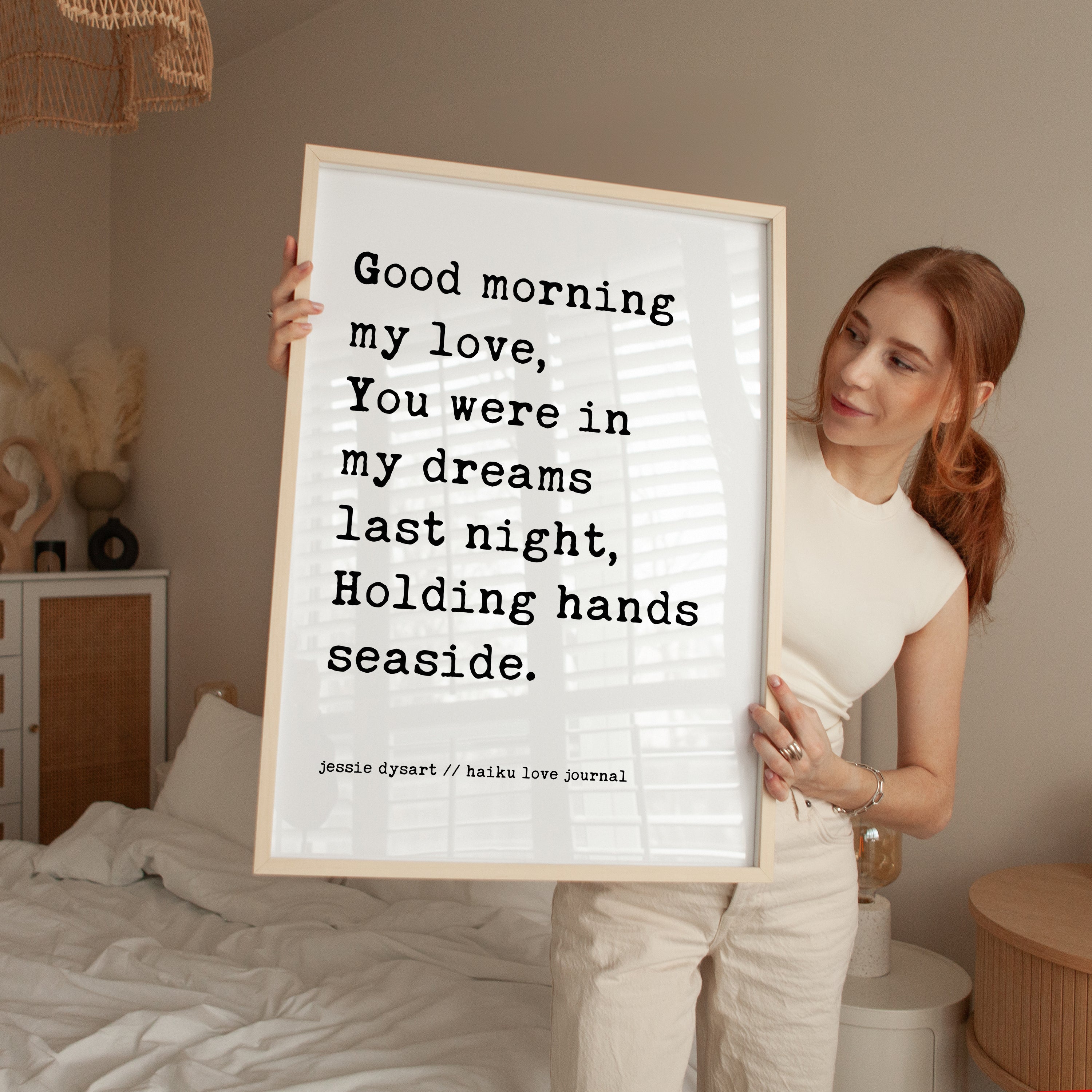 A woman holding a framed poster with a romantic message, standing in a bedroom.