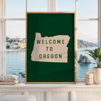 May include: A rectangular wall art print in a light wood frame featuring a dark green background and a beige silhouette of the state of Oregon. The text WELCOME TO OREGON is printed in dark green capital letters centered within the state shape.
