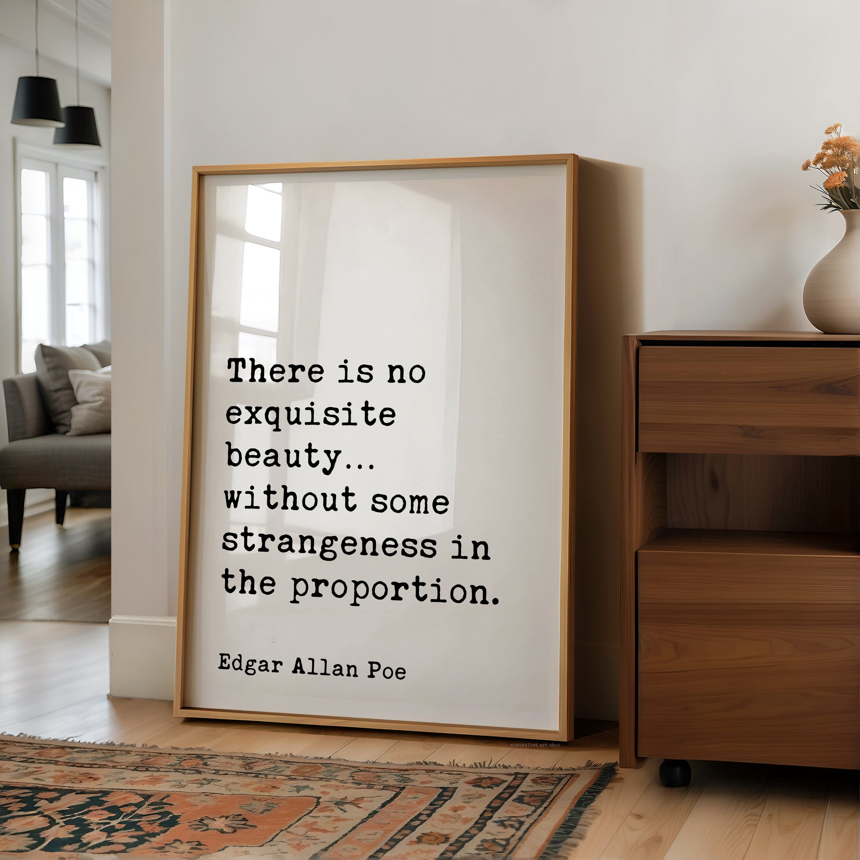 a quote by Edgar Allan Poe, "There is no exquisite beauty... without some strangeness in the proportion." The quote is displayed on a white poster frame, which is placed on a wooden floor in a room with a couch and a vase of flowers.