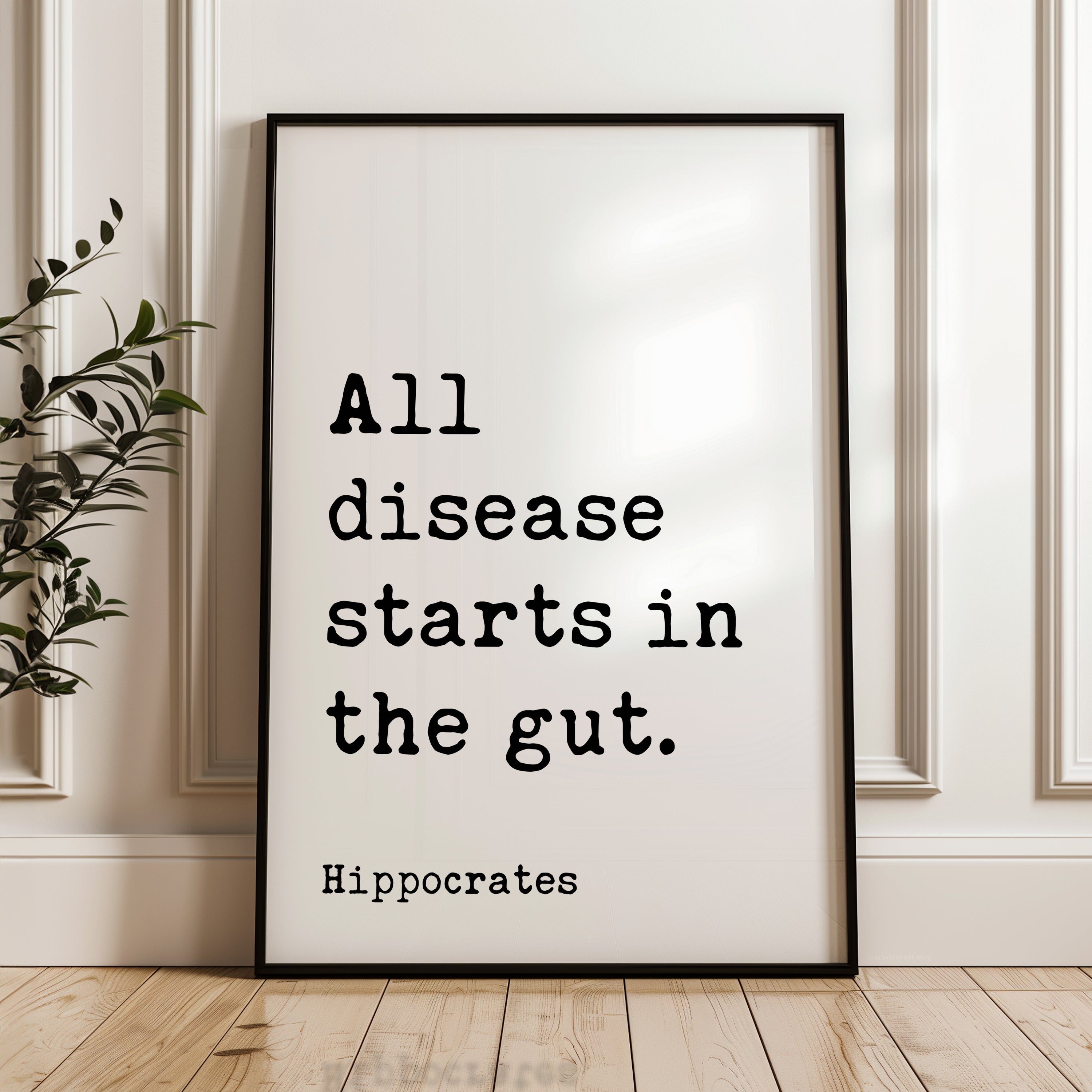 A framed poster with the text "All disease starts in the gut" by Hippocrates, placed on a wooden floor against a white wall.