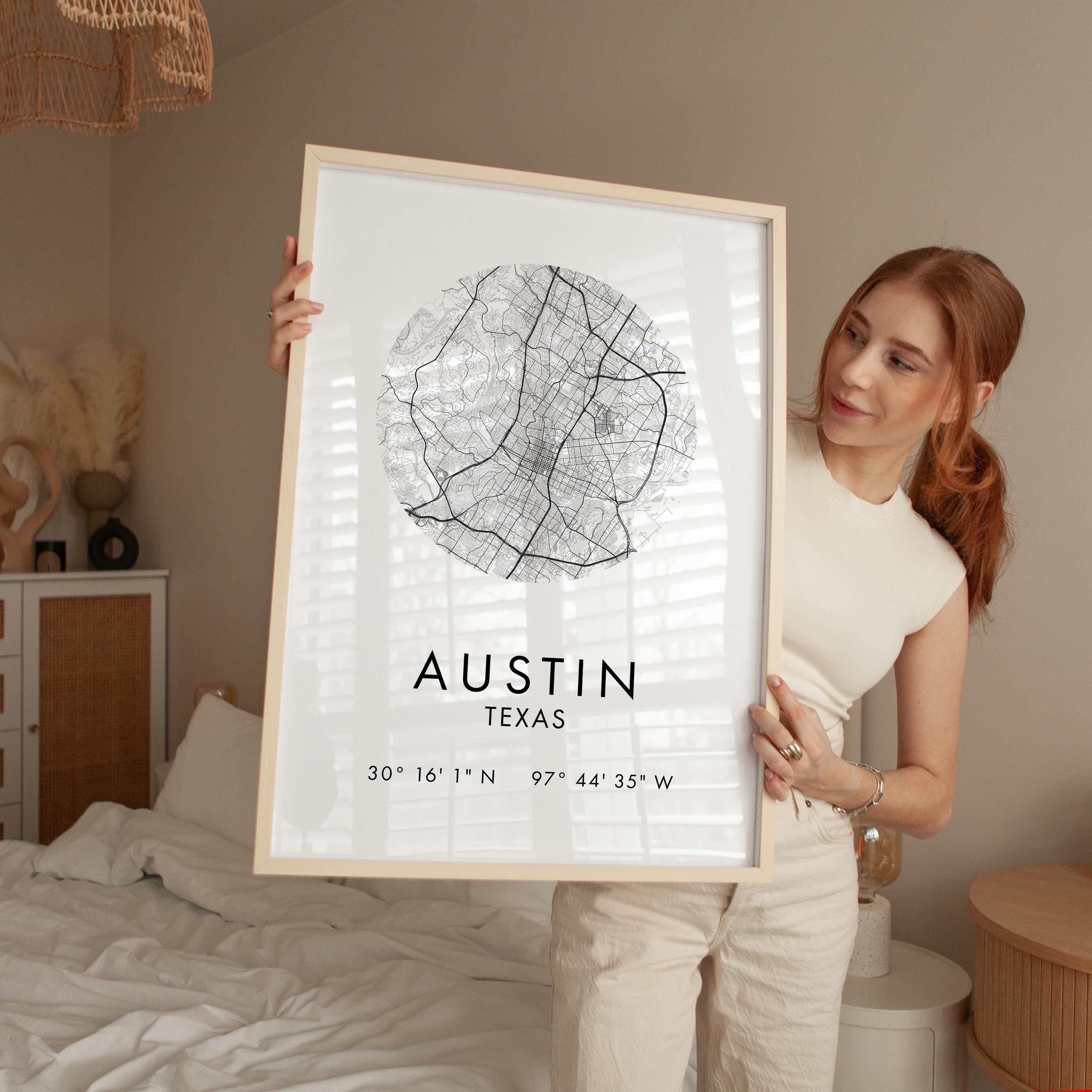 A woman is holding up a framed print of a map of Austin, Texas, with the city's location and coordinates visible.