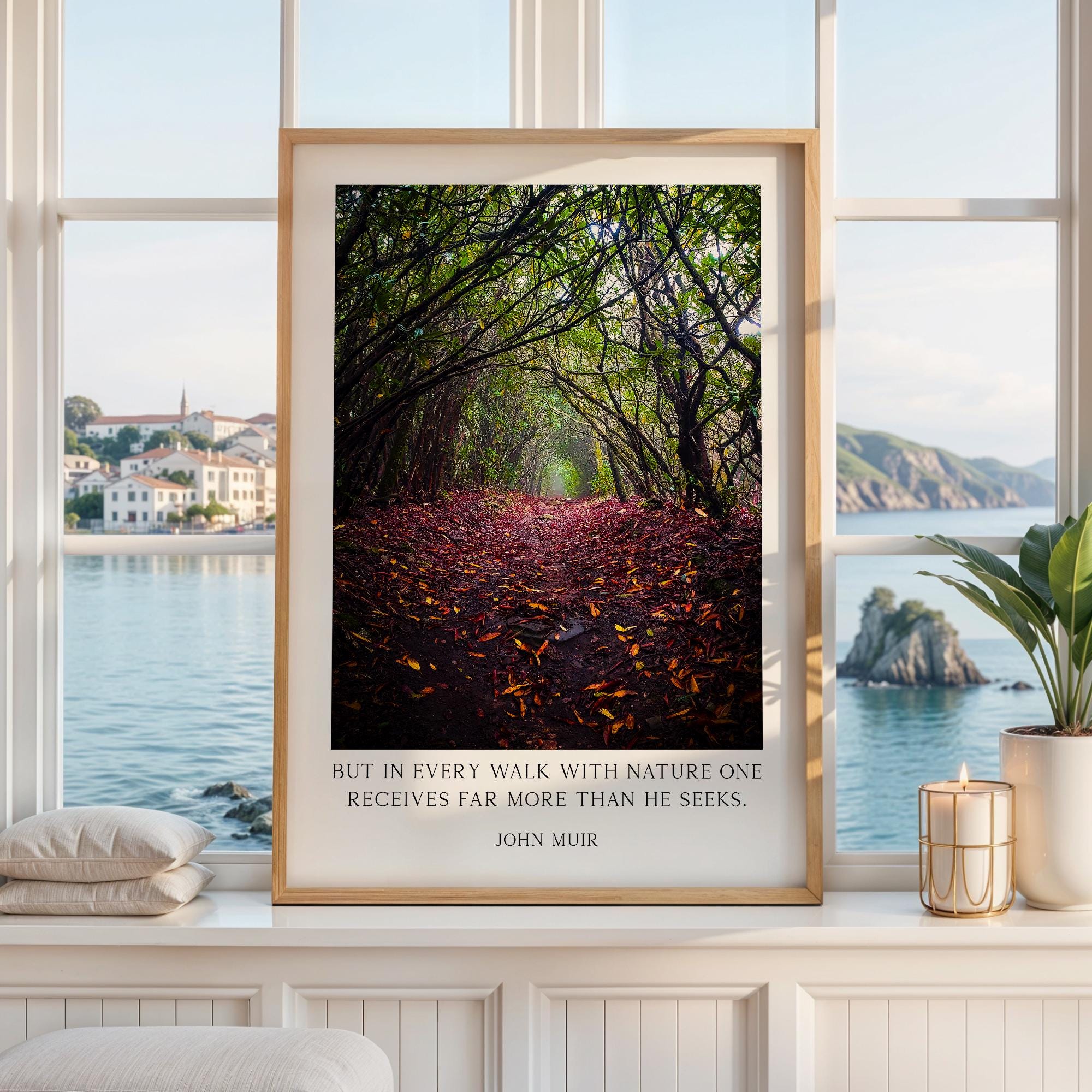 A framed photograph of a forest path with fallen leaves, displayed on a white shelf in a room with a view of a body of water and a distant town.